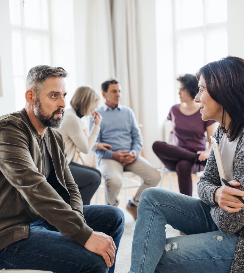 senior-counselor-with-clipboard-talking-to-a-man-during-group-therapy-.jpg